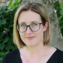 Emma stands in front of a green background with her hair down and wearing glasses and a black top