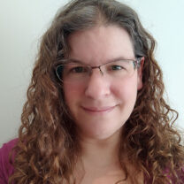 Rachel smiles in front of a light blue background with curly hair and glasses