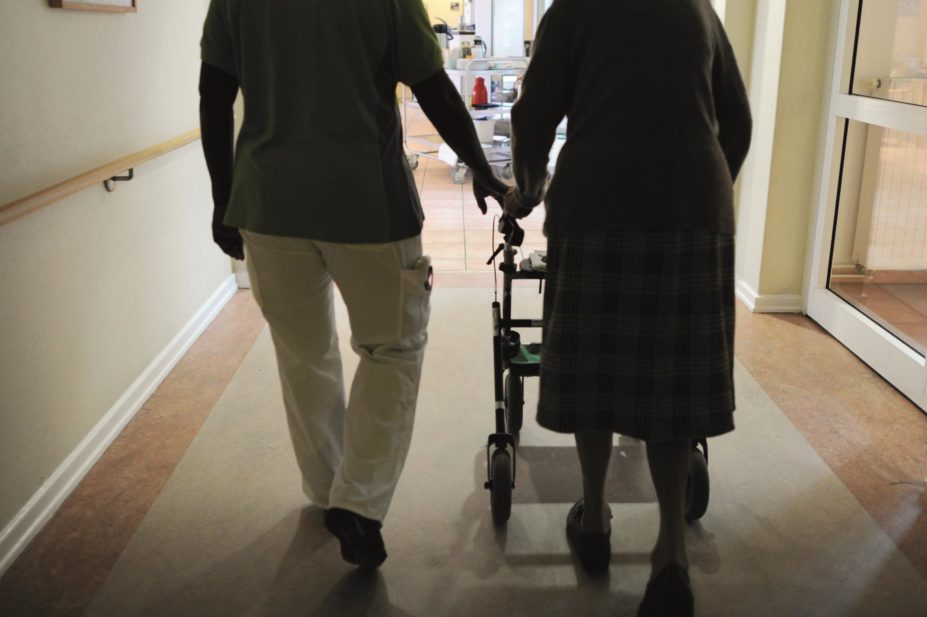 Elderly person on a zimmer frame walking down a corridor of a care home accompanied by a healthcare worker
