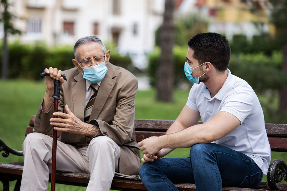 young man wearing mask sat with older man wearing mask holding a walking stick talking on a park bench