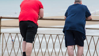 Two obese men leaning on a railing overlooking a beach