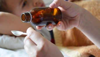 adult pouring medicine into spoon with child lying in background