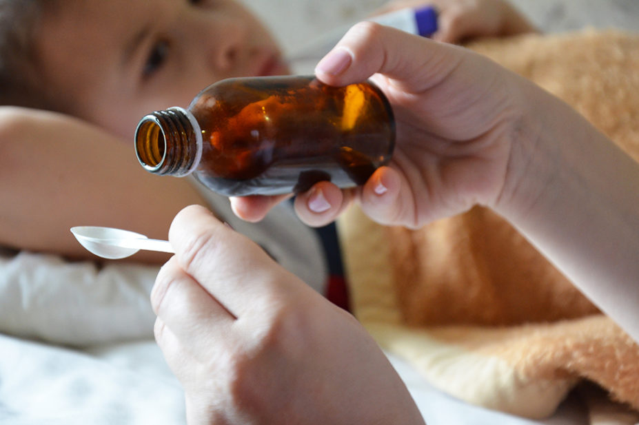 adult pouring medicine into spoon with child lying in background