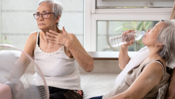 Two older women sit in front of a fan, one drinking bottled water, to avoid the heat.