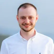 Jonny smiles wearing a white shirt in front of blurred background of sky and grass