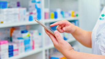 Pharmacist holding computer tablet in front of a pharmacy shelf.