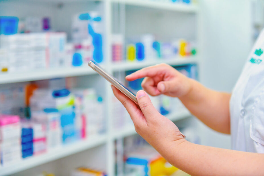 Pharmacist holding computer tablet in front of a pharmacy shelf.