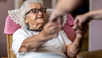 Home carer supporting old woman to stand up from the armchair at care home