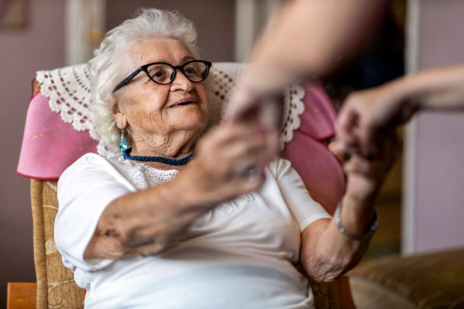 Home carer supporting old woman to stand up from the armchair at care home