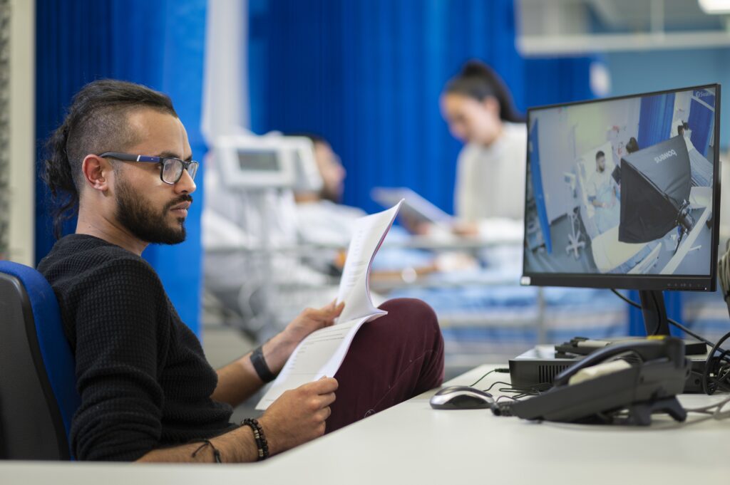 Photo of a tutor looking at a checklist while observing a student on a monitor in a nearby mock clinical environment
