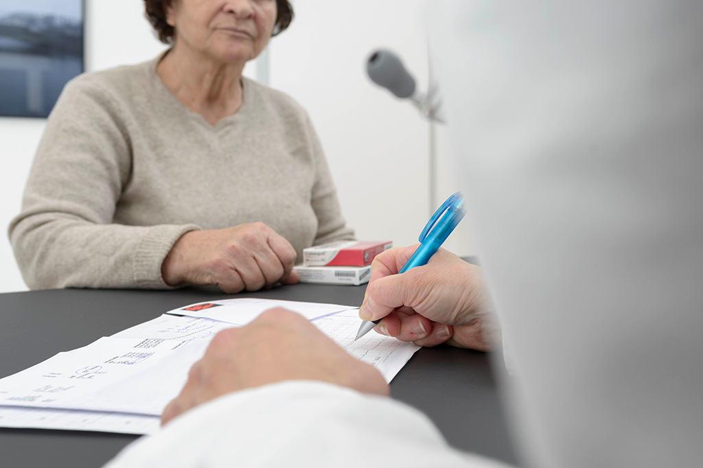 Photo of a hand writing in the foreground, a patient just in focus in the background