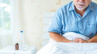 Photo of an older man holding low in his body, sitting up in bed, with water and medication on his bedside table
