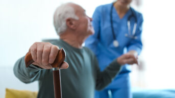Older man with walking stick being helped by physician in his home