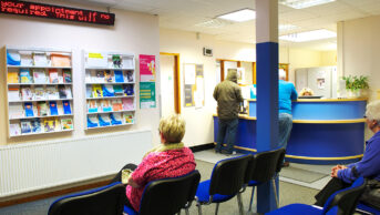 Waiting room in a British GP practice with patients sitting and standing by reception desk waiting to register