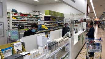 Photo of a customer at a Tesco Pharmacy waiting on a script, with two pharmacists in surgical masks behind the counter