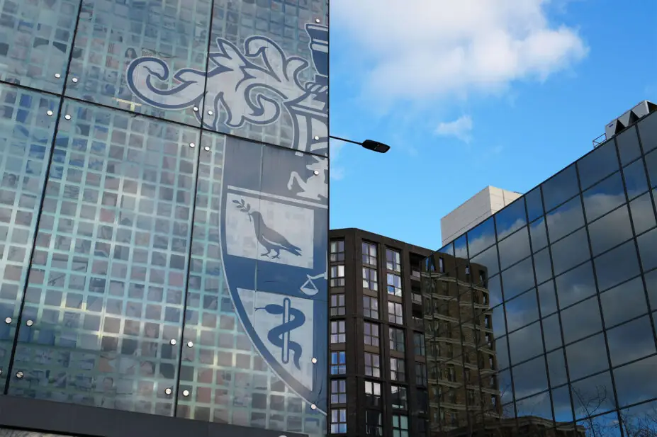 Photo of the outside of the Royal Pharmaceutical Society building on East Smithfield, with half of the RPS logo visible on a blue sky, and other buildings at angles in the background