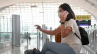 Woman sitting in an airport departures lounge on the phone with a mask on and a band aid on her arm