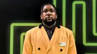 Photo of Emeka Onwudiwe from the waist up, in a yellow overcoat, black shirt and black tie, on a green pharmacy cross background