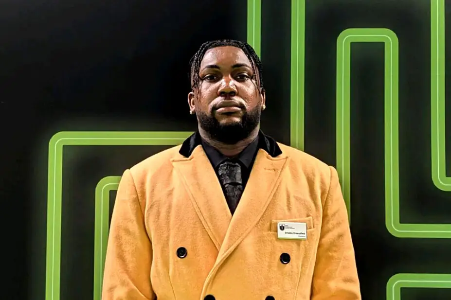 Photo of Emeka Onwudiwe from the waist up, in a yellow overcoat, black shirt and black tie, on a green pharmacy cross background