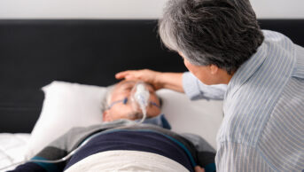 Woman caring for man lying in bed with oxygen mask on, suggestive of palliative care