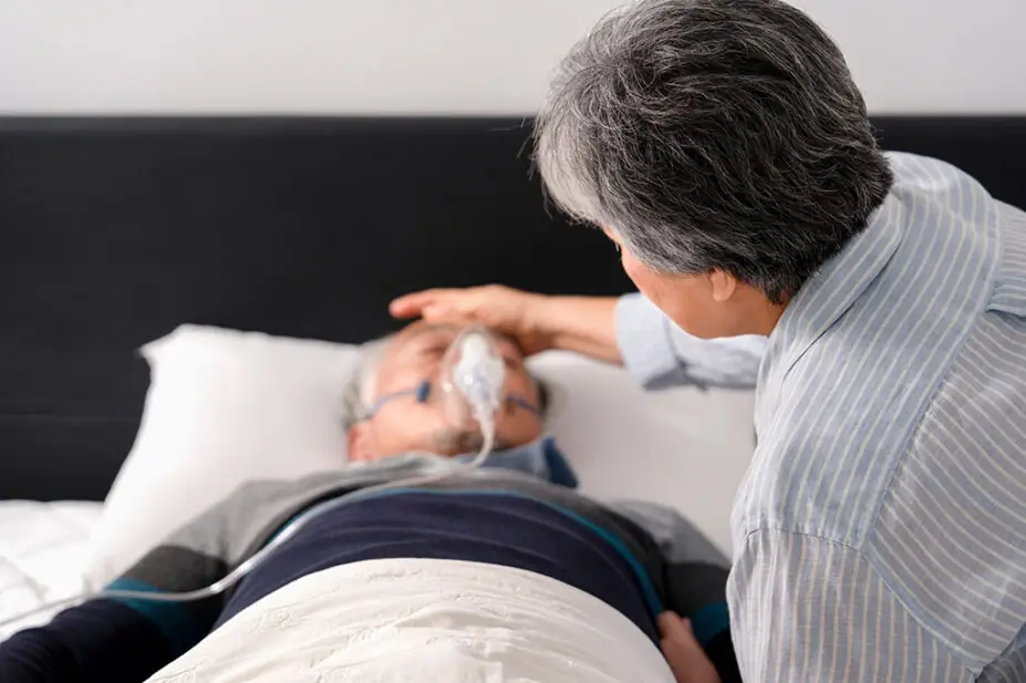 Woman caring for man lying in bed with oxygen mask on, suggestive of palliative care