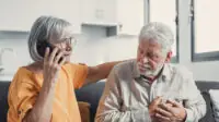 Photo of an older man clutching his chest while a woman calls emergency