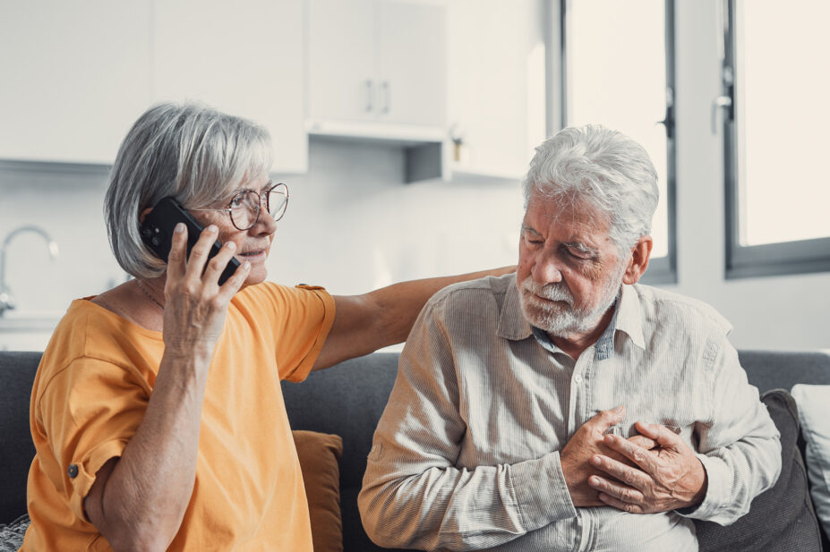 Photo of an older man clutching his chest while a woman calls emergency