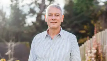 Photo of Bruce Warner in a white and blue vertically-striped collared in a garden, flanked by a fence with autumnal leaves and a soft focused background