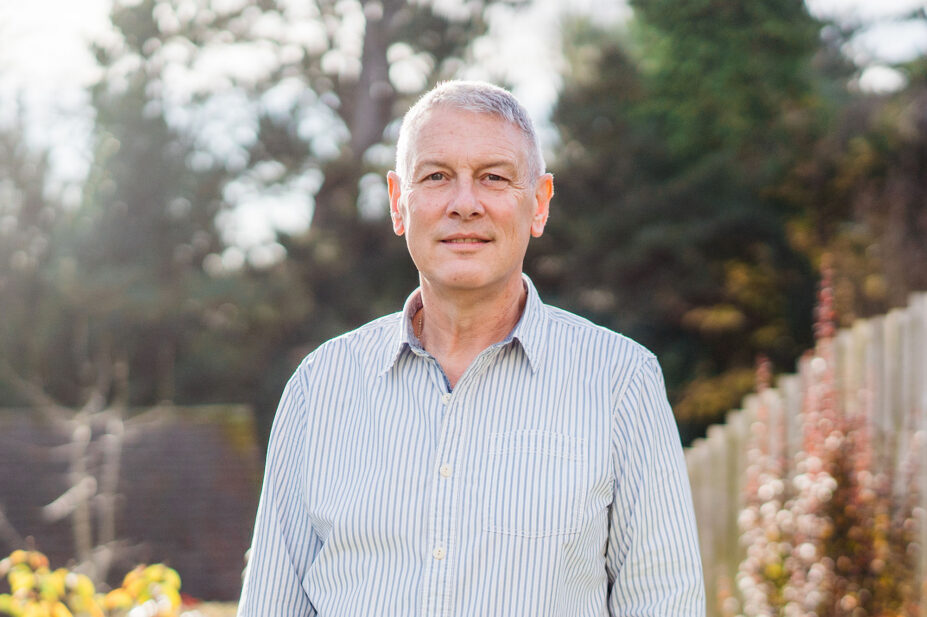 Photo of Bruce Warner in a white and blue vertically-striped collared in a garden, flanked by a fence with autumnal leaves and a soft focused background