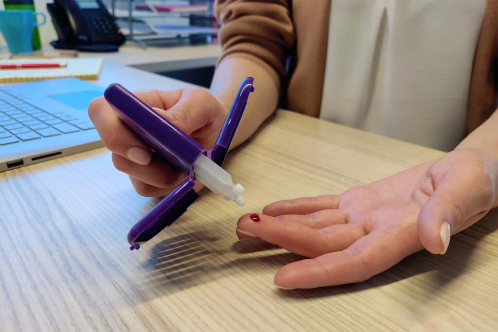 Photo of a hand with a pin prick blood drop on the middle finger, with the person's other hand hovering over top holding a purple device with a collection dropped and winged pieces either side