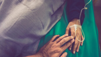 Someone holding older person's hands in sick bed