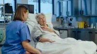 Photo of older woman in hospital bed who looks in pain, talking to a nurse