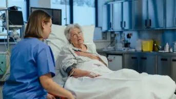 Photo of older woman in hospital bed who looks in pain, talking to a nurse