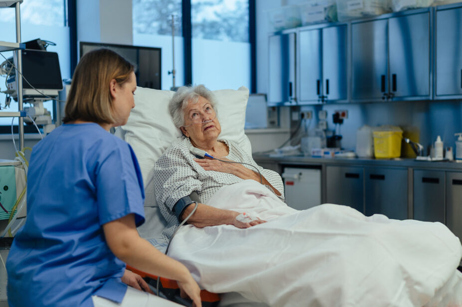 Photo of older woman in hospital bed who looks in pain, talking to a nurse