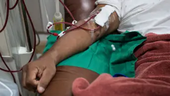 Photo of the arm of a dark-skinned man receiving haemodialysis on a dark brown couch, with a burgundy blanket over his knees