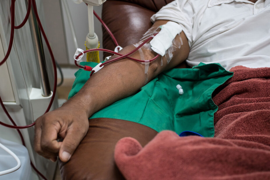 Photo of the arm of a dark-skinned man receiving haemodialysis on a dark brown couch, with a burgundy blanket over his knees