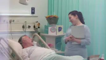 Photo of a nurse or pharmacist in hospital holding a clipboard and looking down with care at a patient in bed