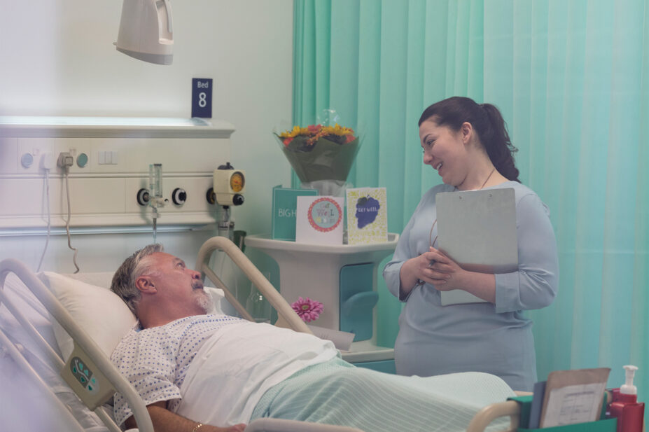 Photo of a nurse or pharmacist in hospital holding a clipboard and looking down with care at a patient in bed
