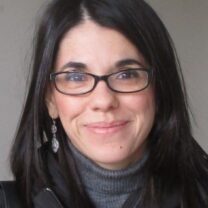 Olga smiles at the camera wearing black glasses in front of a neutral grey background