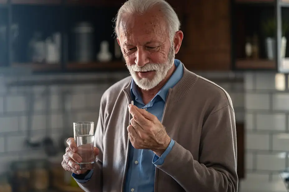 An older man takes a white pill with a glass of water