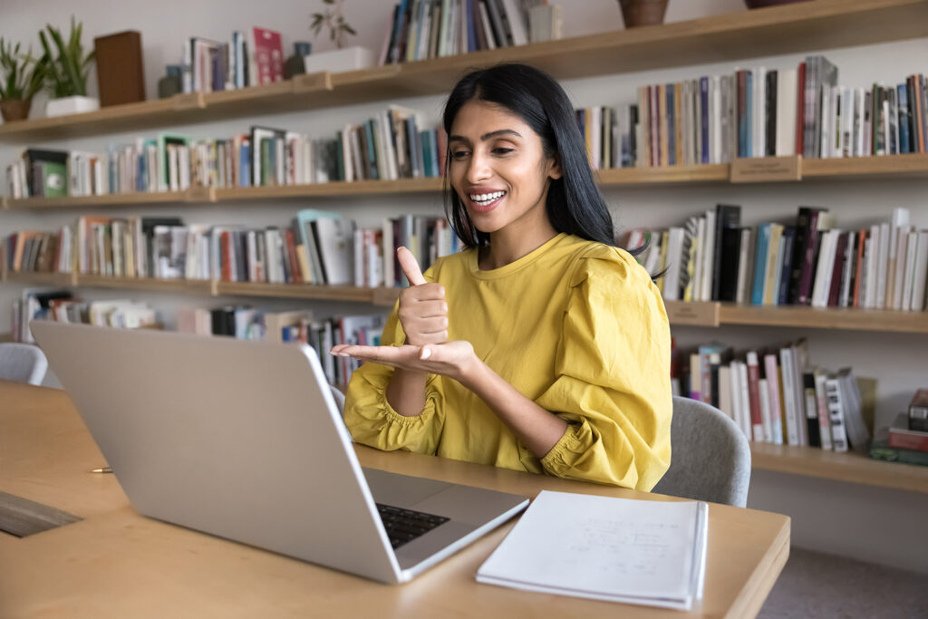 Woman speaking to student with hearing disability, showing hand gesture at laptop, working at computer in library