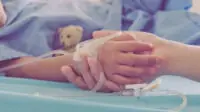 An adult holds the hand of a child patient in a hospital bed