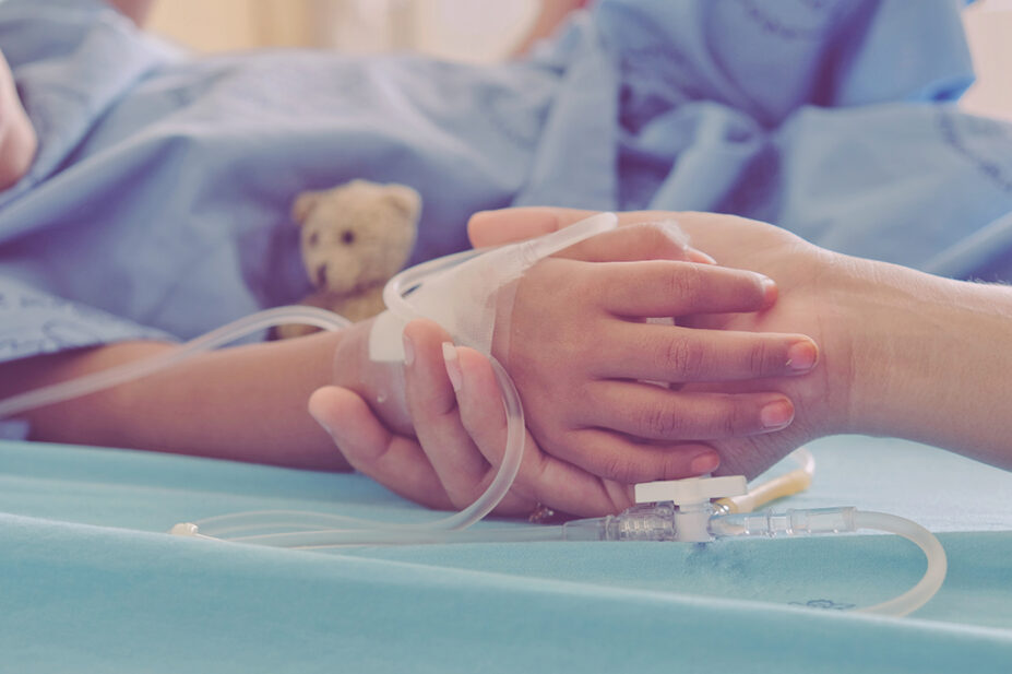 An adult holds the hand of a child patient in a hospital bed