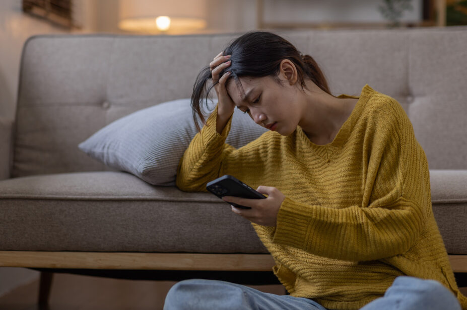 A woman on her phone sits on the floor by the sofa, looking worn out and depressed