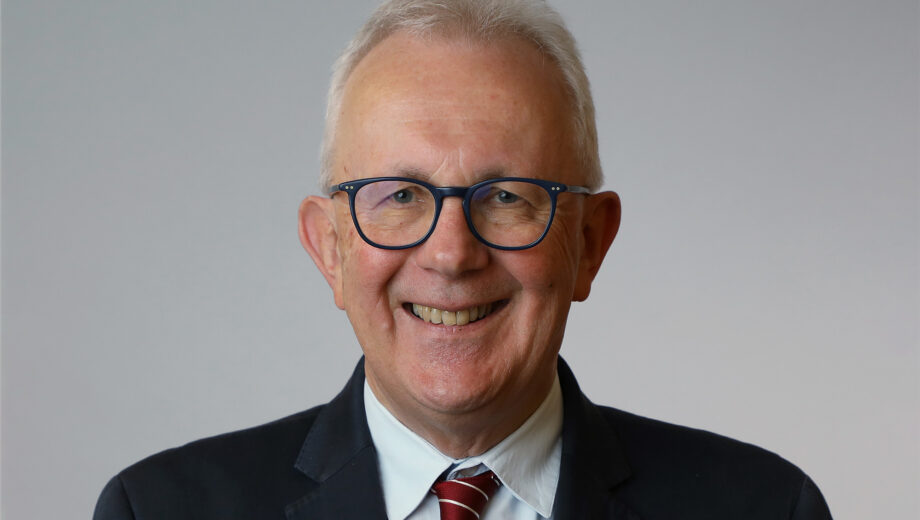 Hugh stands in front of a grey background, he is smiling, wearing a black suit and red tie