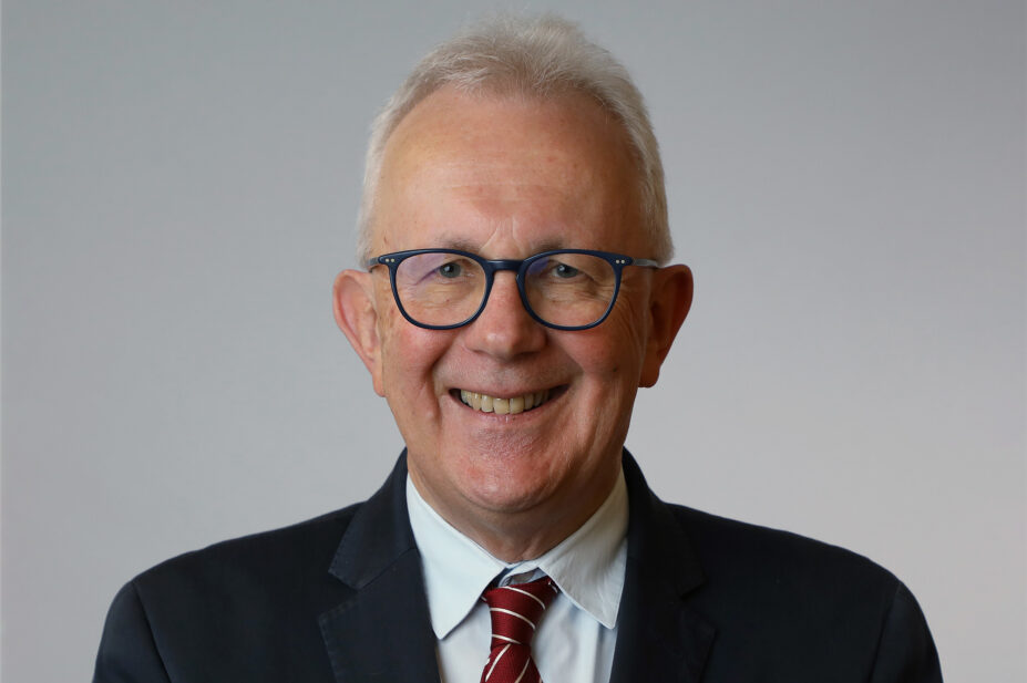 Hugh stands in front of a grey background, he is smiling, wearing a black suit and red tie