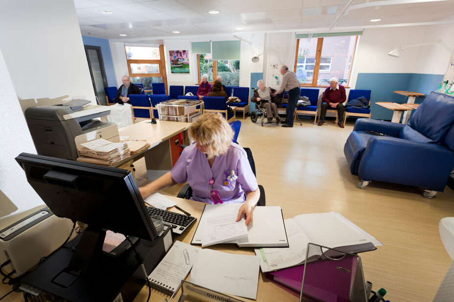 Person at desk with lots of papers and computer, lots of people behind in waiting area