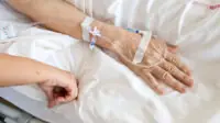 A older patient with a catheter inserted in their hand lays in a hospital bed