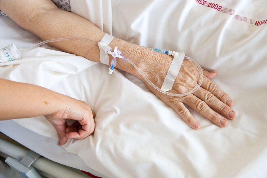 A older patient with a catheter inserted in their hand lays in a hospital bed
