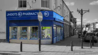 The outside of a Jhoots pharmacy on a high street. Background is in black and white